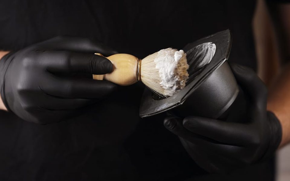 Close up photo of hairstylist hands holding bowl of shaving foam and wooden shaving brush. man He in black glovers is standing indoor barbershop . Shaving accessories